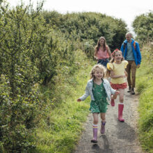 A medium wide angle view of a family of four exploring the coastal trails along the coast on a beautiful sunny summers day in Craster in the North East of England. The two daughters are running ahead of their parents and following the path.